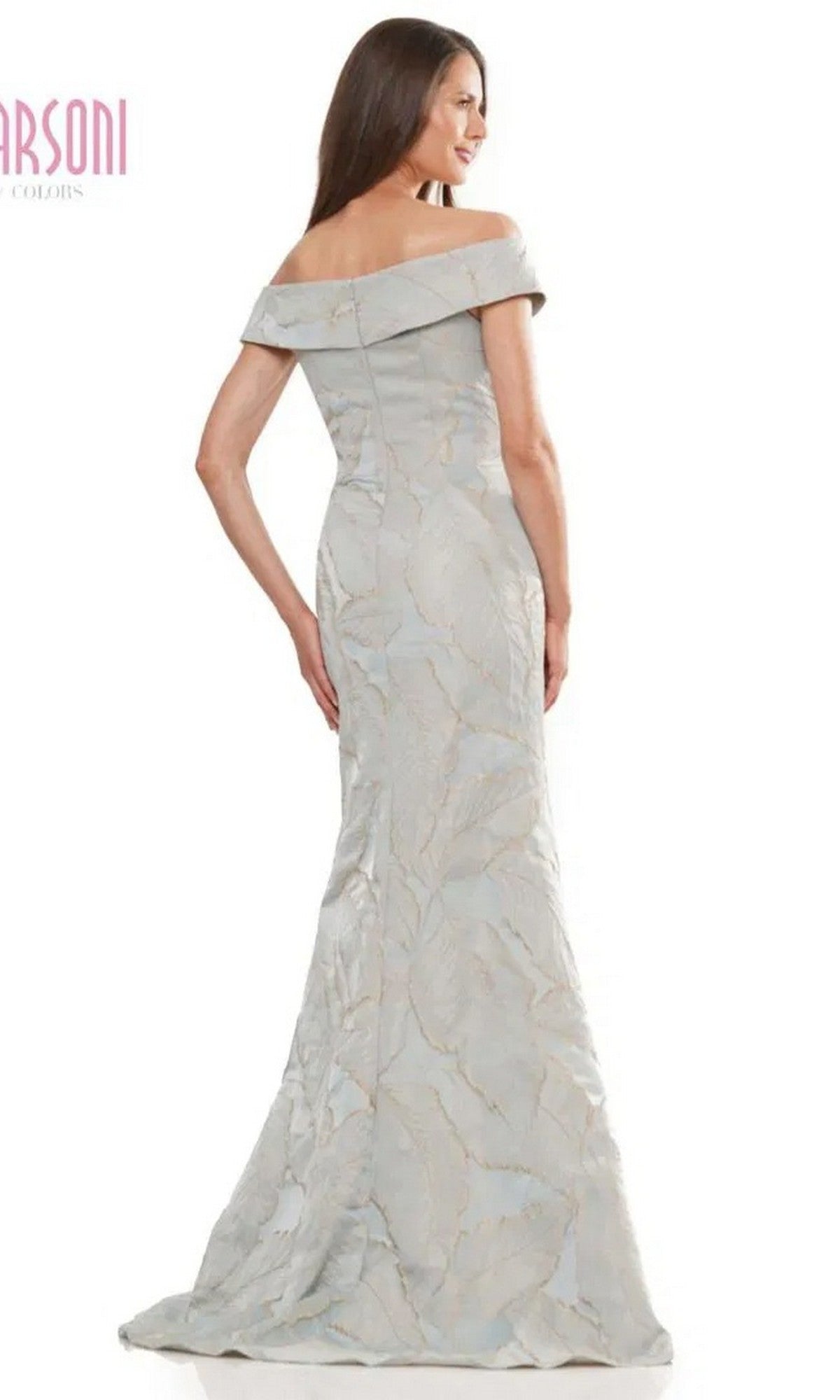 A woman stands facing away from the camera in the Marsoni MV1266, a long formal light grey off-the-shoulder evening gown featuring a subtle leaf pattern.